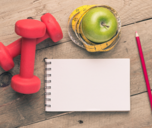 Red weights, green apple, measuring tape, paper, and pencil on table.